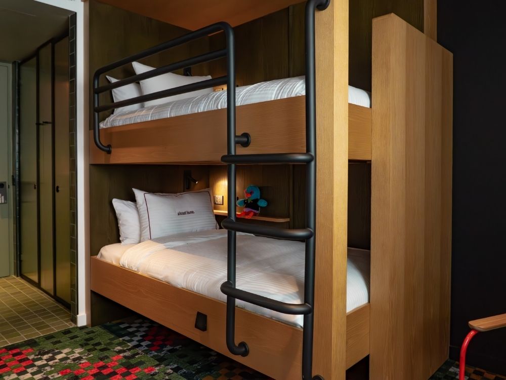 Wooden bunk beds with white linens and pillows in a modern room featuring a decorative ladder and colorful checkered carpet.