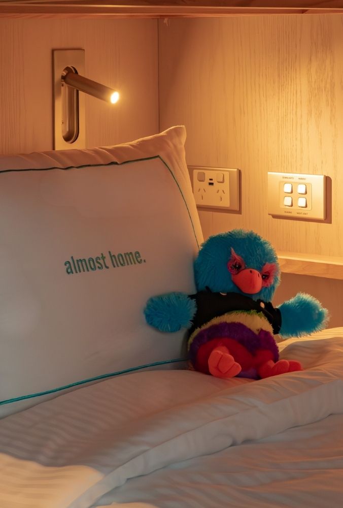Cosy bedroom corner with a pillow reading "almost home" and a colorful plush bird on a neatly made bed under a reading light.
