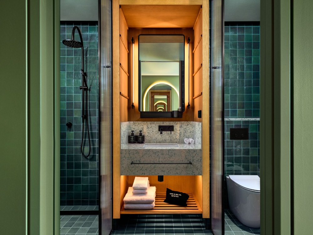 Stylish bathroom featuring teal tiles, a marble sink, shelves with towels, a round mirror, and a gold shower fixture.