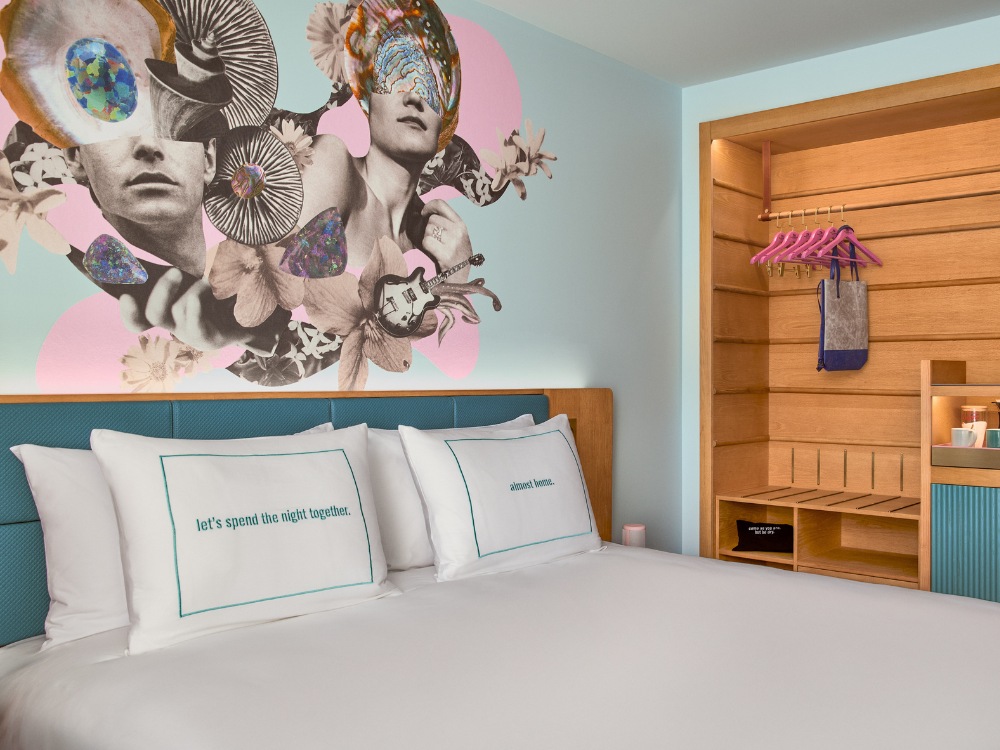 A stylish hotel room featuring a vibrant mural, a cozy bed with decorative pillows, and a wooden wardrobe with colorful hangers.