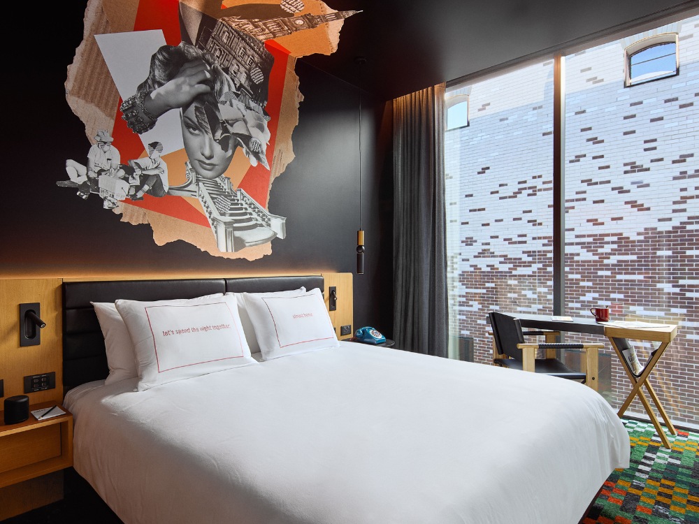 A modern hotel room featuring a king-sized bed, artistic wall decor, a workspace with a chair, and large windows overlooking a stylish exterior.