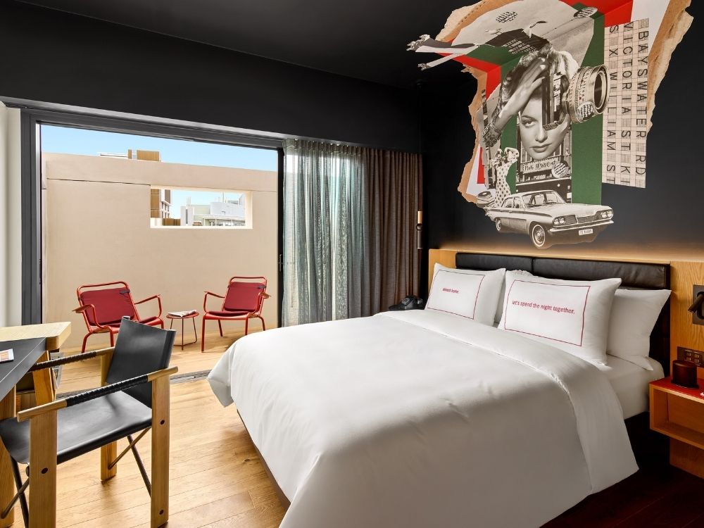 Modern hotel room with large bed, artistic collage above headboard, sliding glass door opening to balcony with red chairs and small table.