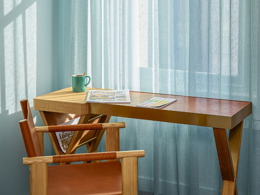 A stylish workspace features a wooden table with a coffee mug and magazines, alongside a modern chair and sheer blue curtains.