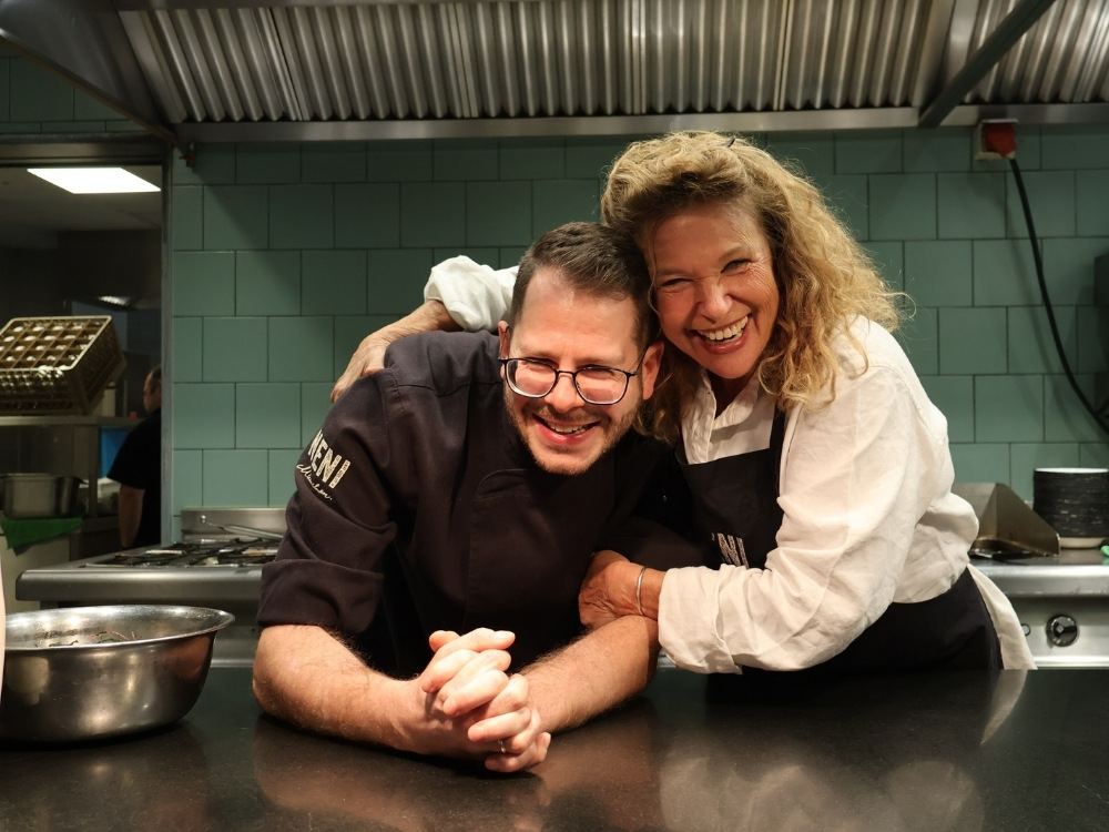 A smiling man in a chef's coat and glasses leans on a kitchen counter, embraced by a cheerful woman in a white shirt. They're in a restaurant kitchen.