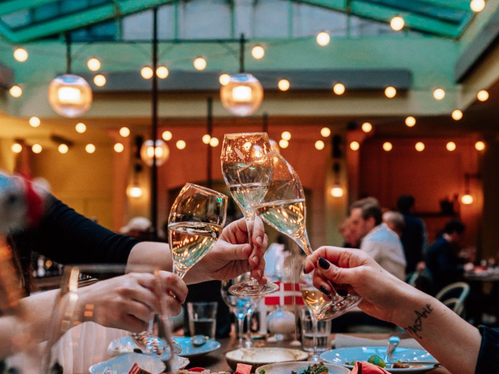 Alt text: Guests clinking glasses of white wine over a shared dinner table at NENI Copenhagen, surrounded by warm lights and a cozy indoor setting.