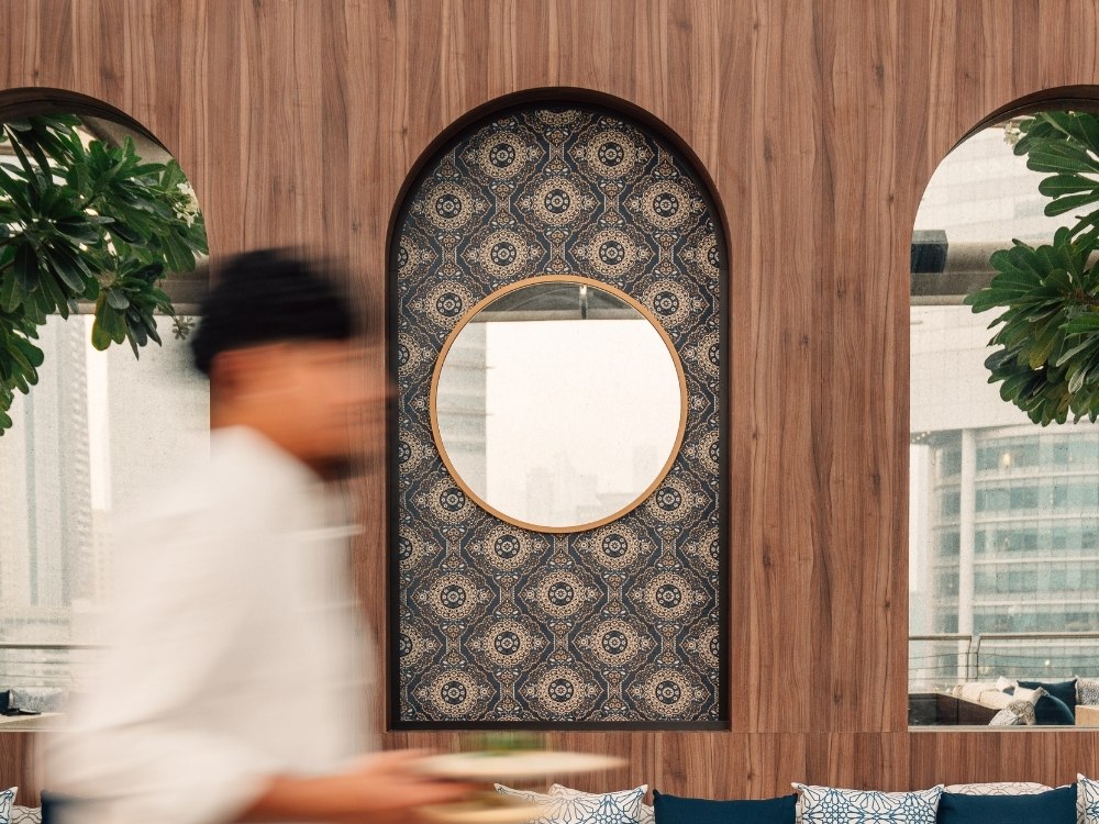 Blurred figure in white passes a wooden wall with an ornate, circular mirror. Green plants frame the archways, creating an elegant ambiance.