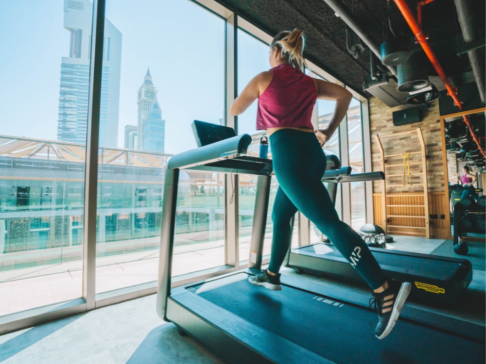 A woman in athletic wear runs on a treadmill in a modern gym. Large windows reveal a cityscape featuring tall buildings. The mood is energetic and focused.