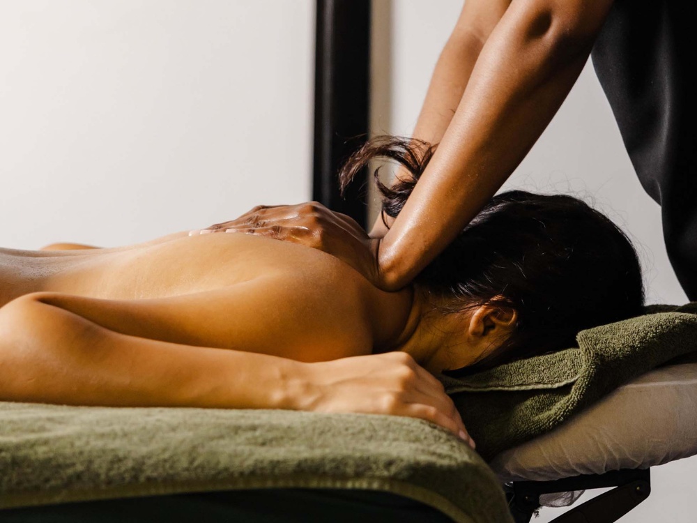 A woman is lying face down on a massage table with a green towel, receiving a back and neck massage. The scene conveys relaxation and comfort.