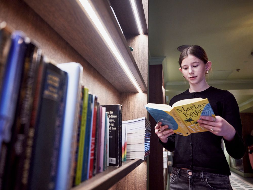 A young woman reading a book while standing next to a shelf filled with books.
