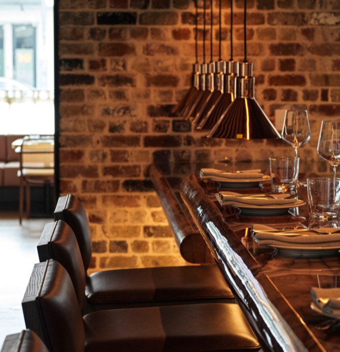 A stylish dining area featuring a wooden table set with plates and glasses, illuminated by modern pendant lights against a brick wall.