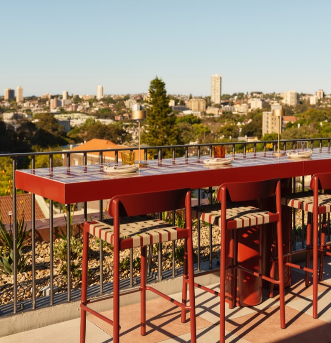 Rooftop bar with red table and striped stools overlooking a city skyline, bathed in warm sunlight.