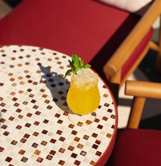 A vibrant yellow cocktail with crushed ice and a mint sprig sits on a tiled table, surrounded by red upholstery.