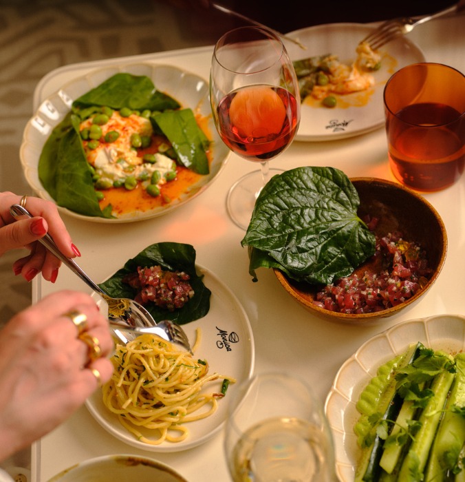 A vibrant table setting featuring various dishes: pasta, fresh vegetables, and a glass of rosé wine, with hands engaged in dining.