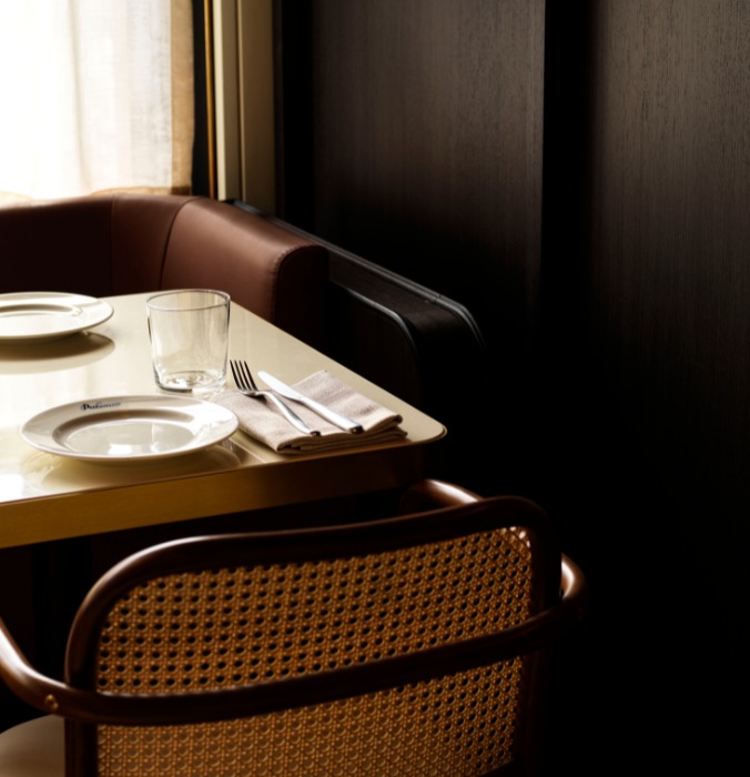 A cosy dining table set with plates, a glass, and utensils, adjacent to a plush, brown booth in a dimly lit restaurant.