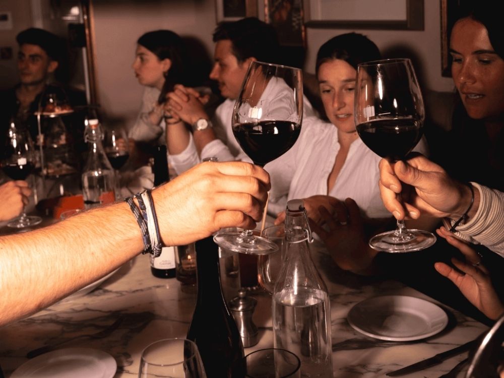 A lively dinner party scene with people around a marble table, raising glasses of red wine. The mood is celebratory and warm, with dim lighting.