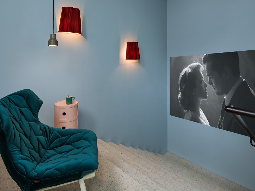 A cozy room with a teal cushioned chair, pink side table, and two red-shaded wall lights. A black-and-white romantic movie scene is projected on a blue wall.