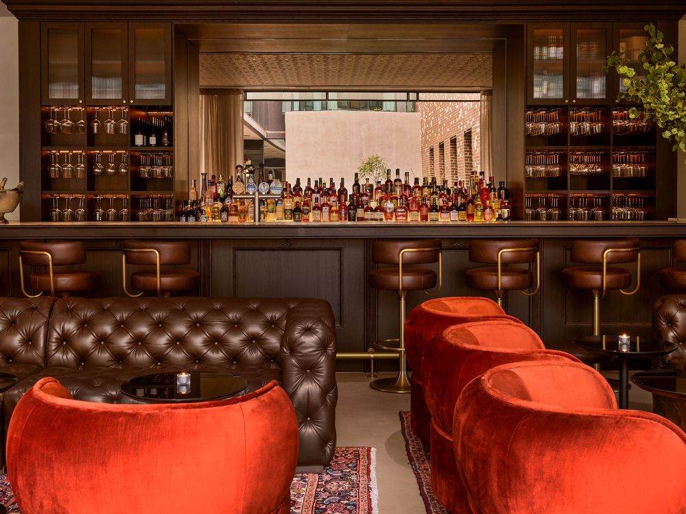 Elegant bar with a warm, inviting atmosphere. Features plush red chairs, brown leather sofas, a stocked bar with liquor bottles, and cosy lighting.