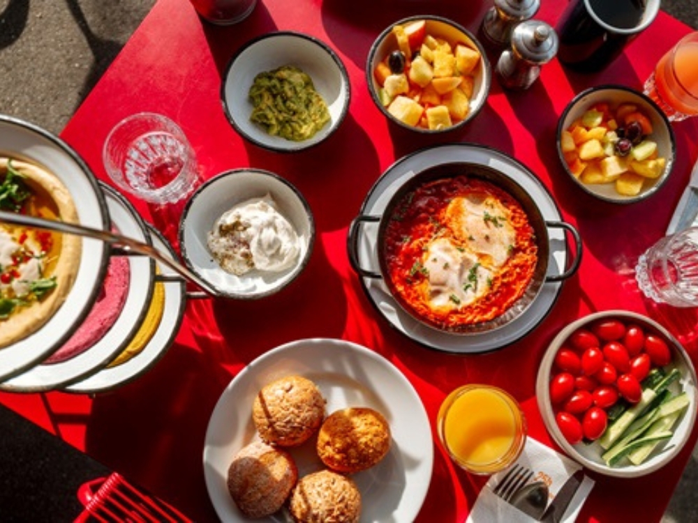 A vibrant brunch spread on a red table, featuring shakshuka, fresh fruits, yogurt, muffins, cherry tomatoes, and drinks.