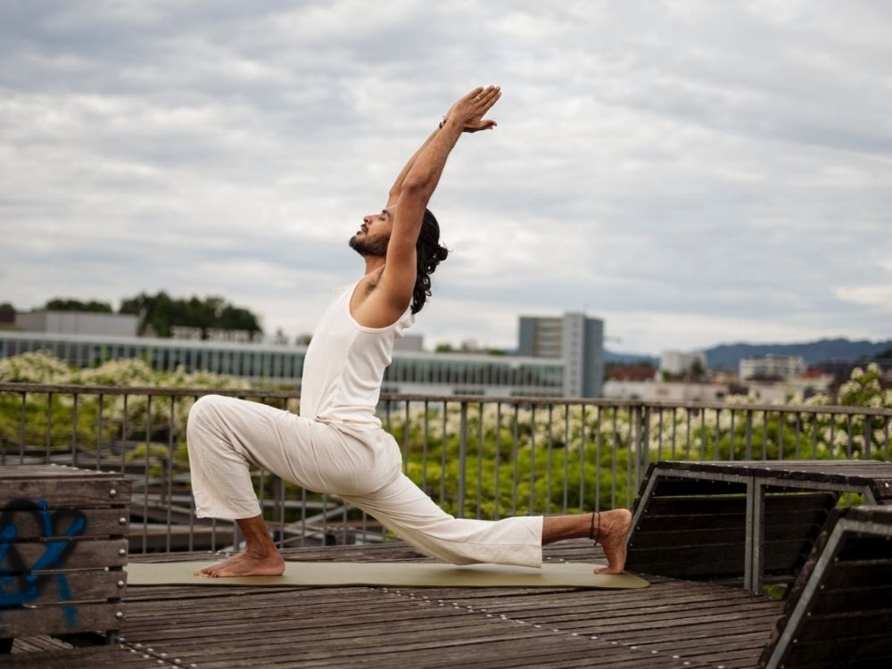 A person practices yoga in a high lunge pose on a rooftop against a cloudy sky, wearing white clothes.