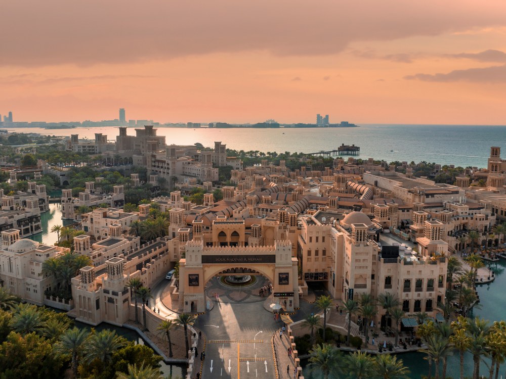 Aerial view of a stunning resort city by the sea, showcasing intricate architecture and palm-lined walkways under a colorful sunset sky.