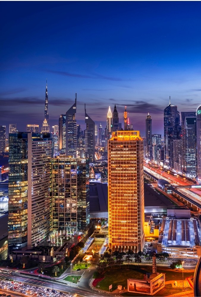 Dubai skyline at dusk, featuring brightly lit skyscrapers with glowing lights. A busy highway with streaks of light from vehicles adds energy to the scene.