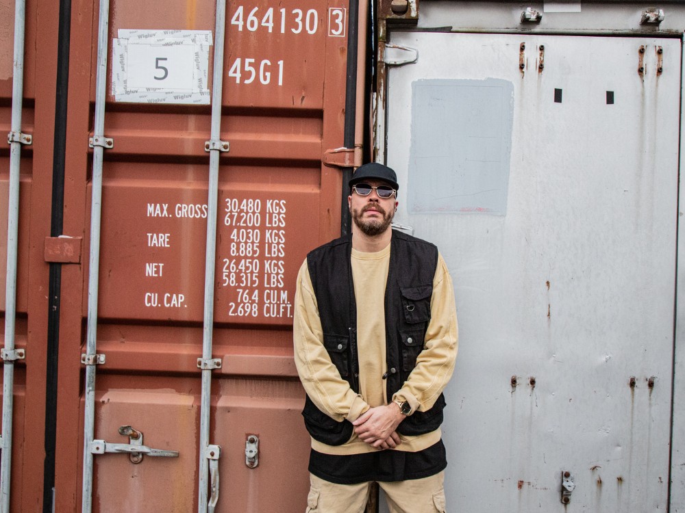 A person wearing a beige shirt and black vest stands in front of a shipping container, displaying cargo specifications and weights.