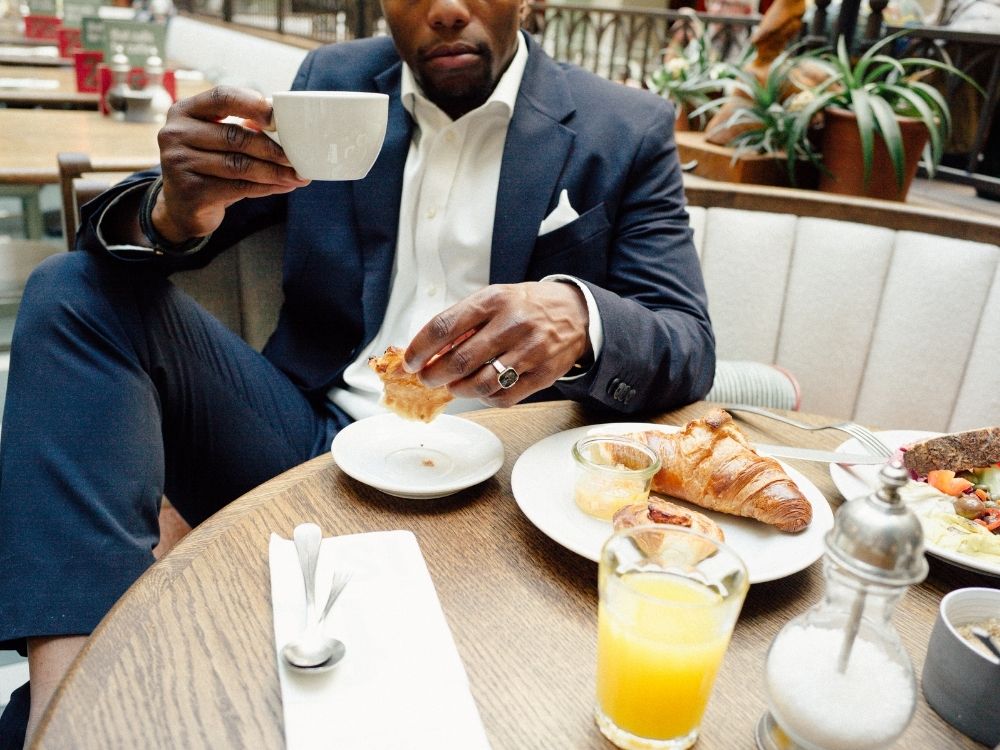 A man in a navy suit sits at a café table, holding a cup of coffee and eating a pastry. On the table are a croissant, small jars of jam, orange juice, cutlery, and a plate of breakfast items.