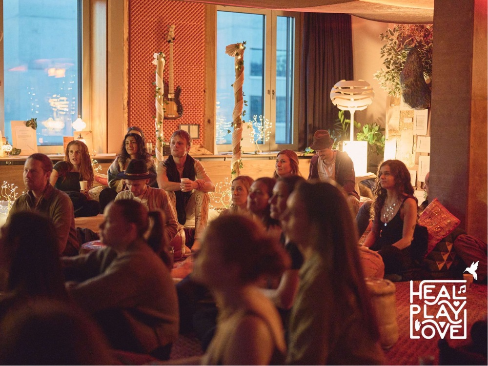 A cozy gathering of people sits attentively on the floor, illuminated by ambient lighting and fairy lights. The room's warm, intimate atmosphere is enhanced by plants, patterned textiles, and musical instruments, conveying a sense of relaxation and focus.