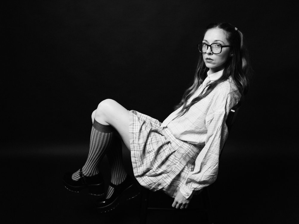Black and white photo of a young woman in glasses, sitting on a chair. She wears a plaid skirt, knee-high socks, and looks thoughtfully to the side.