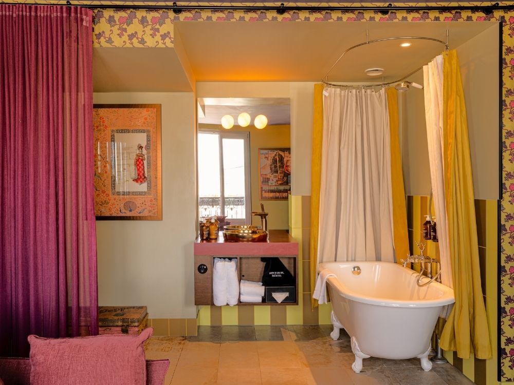 A charming bathroom with a vintage style features a freestanding white bathtub with yellow and white curtains. The walls are adorned with decorative patterns and framed art.