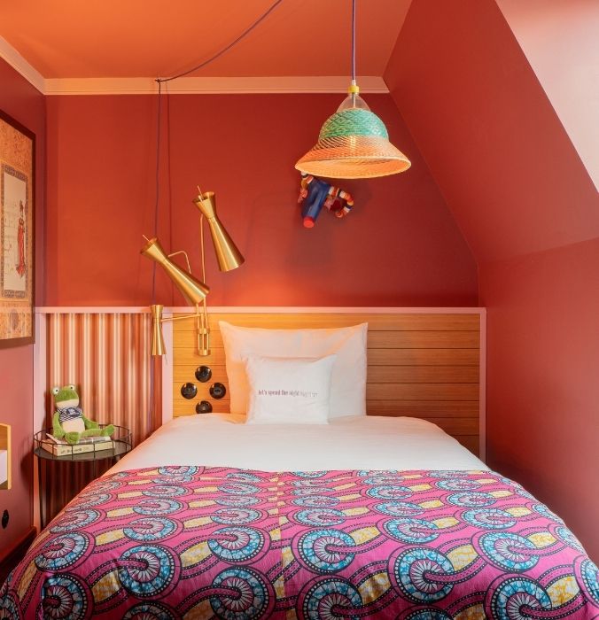 Cosy bedroom with a single bed against a warm terracotta wall. The bed has a colorful patterned quilt, white pillow, and wooden headboard.