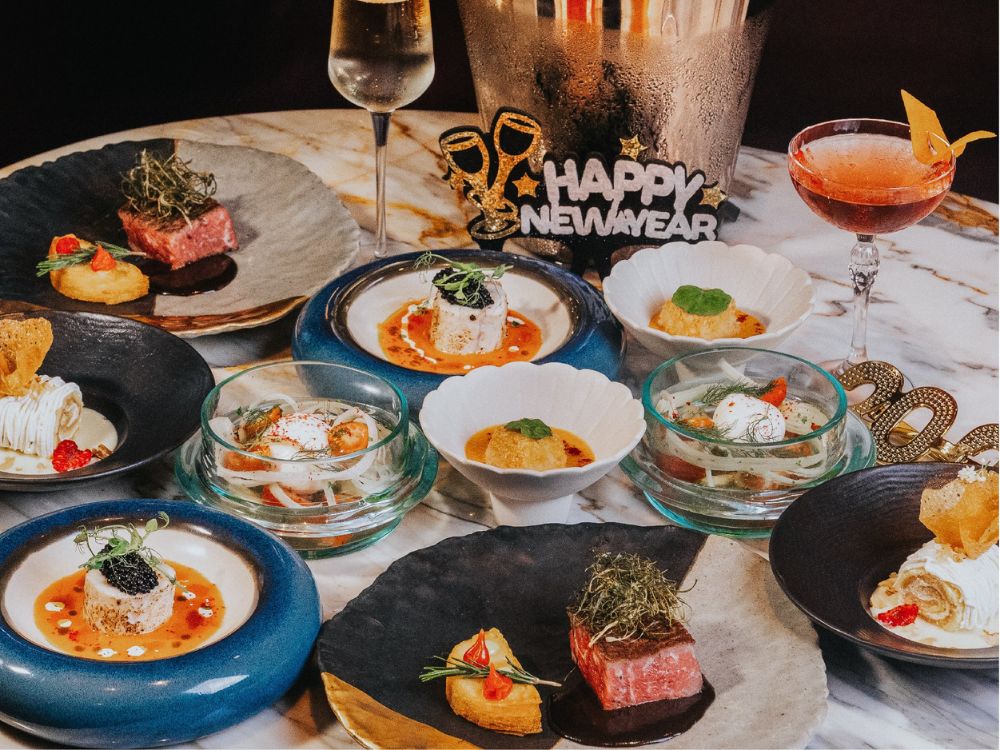 Elegant New Year's table with artful dishes on black and blue plates, festive "Happy New Year" sign, champagne, and cocktails, exuding celebration.