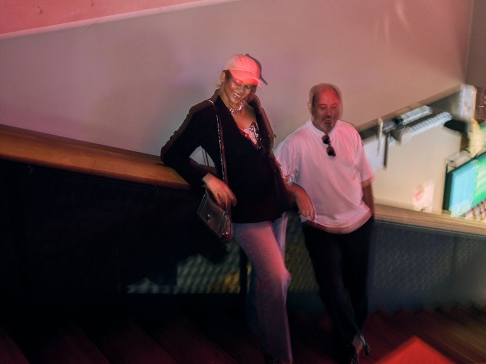 A woman in a pink cap leans on a stair railing, smiling. She wears a black jacket and jeans. Beside her, a man in a white shirt stands casually. Dim lighting suggests a relaxed, informal setting.