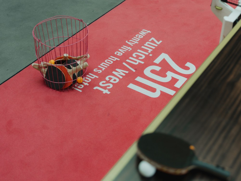 A red table tennis court floor with a wire basket holding paddles and balls. Text reads "25h zurich/west twenty five hours hotel." One paddle and a ball lie nearby.