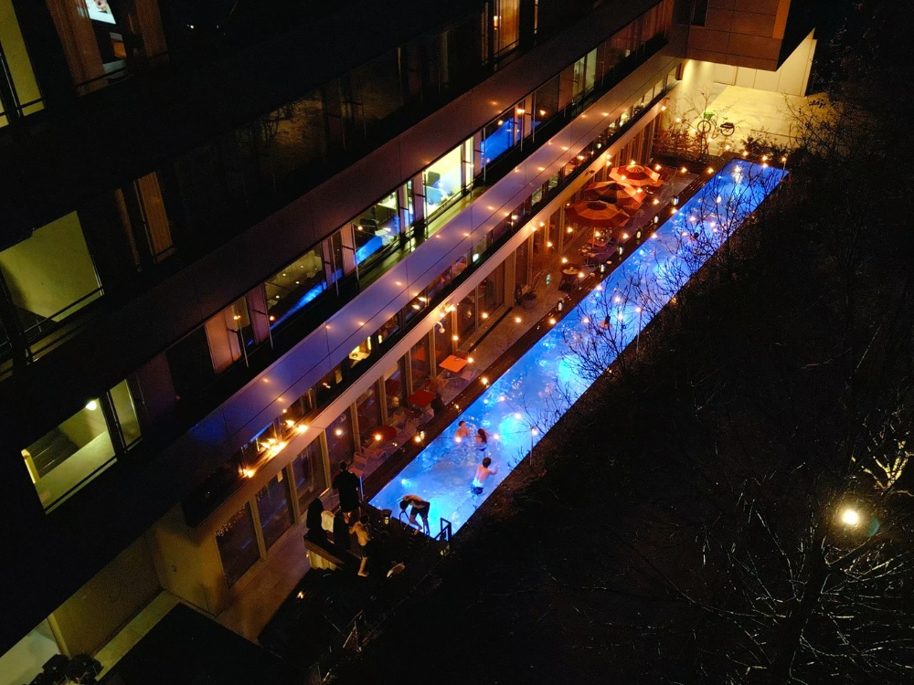 Aerial view of a hotel with a glowing blue pool lined by string lights. People swim and relax at tables under umbrellas, creating a warm, lively atmosphere.
