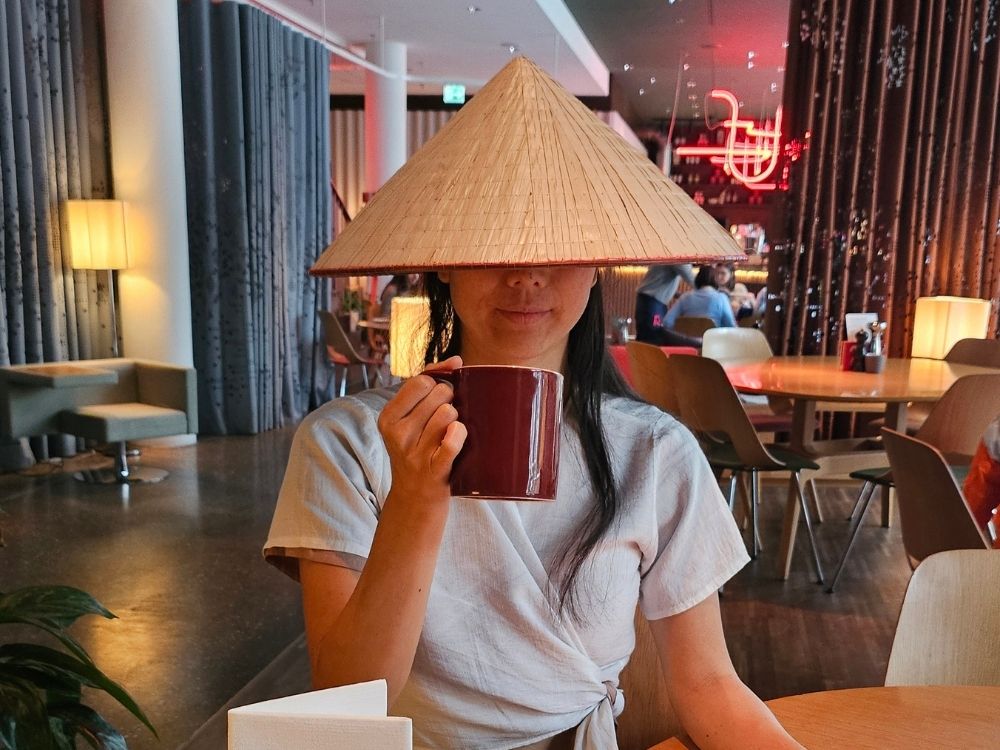 Person wearing a conical hat, sipping from a large mug in a modern café. The ambiance is cozy with warm lighting. The scene is playful and relaxed.