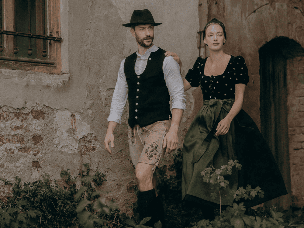 A man and woman in traditional Bavarian attire walk by an old stone building. The scene feels rustic and nostalgic with earthy tones.
