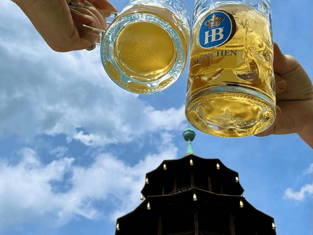 Hands raise beer mugs against a blue sky with scattered clouds. Below, a dark pagoda-style tower creates a celebratory, leisurely atmosphere.