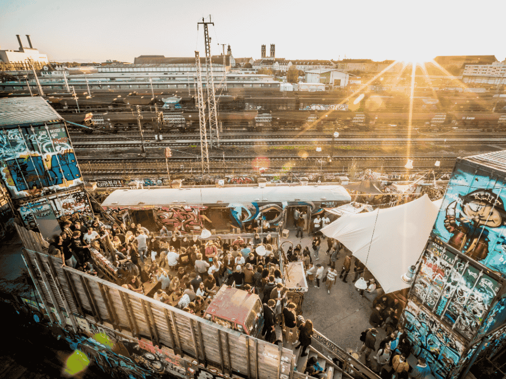 A lively outdoor gathering beside graffitied train tracks at sunset. People socialize amid vibrant street art, with bright sun rays casting a warm glow.