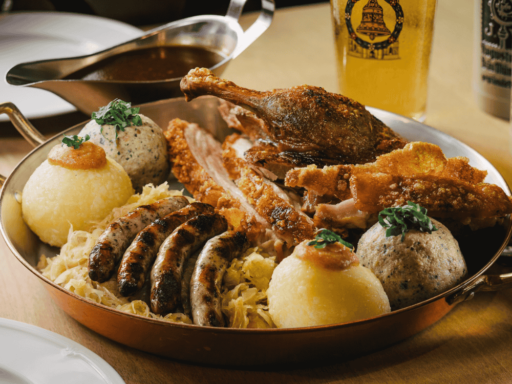 A hearty platter featuring crispy roast duck, sausages, dumplings, and sauerkraut on a wooden table, accompanied by a glass of beer.