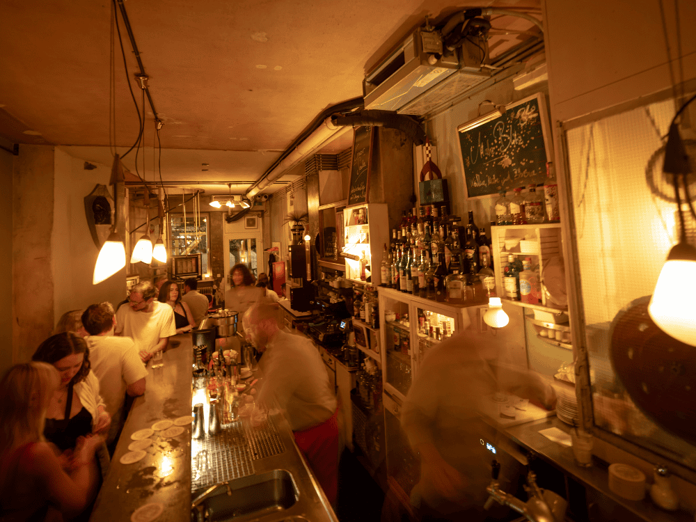 Warmly lit bar with patrons chatting at the counter. Shelves of bottles and hanging lights create a cozy, bustling atmosphere.