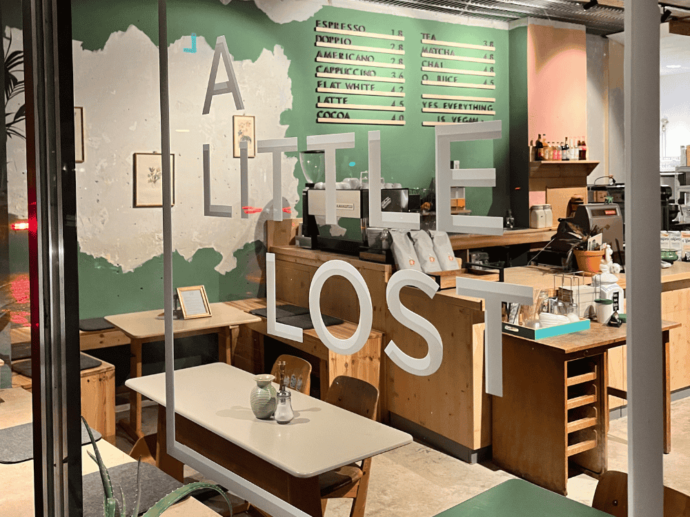 Cosy cafe interior with a wooden counter, warm lighting, and green walls. The phrase "A Little Lost" is on the window, creating a welcoming vibe.