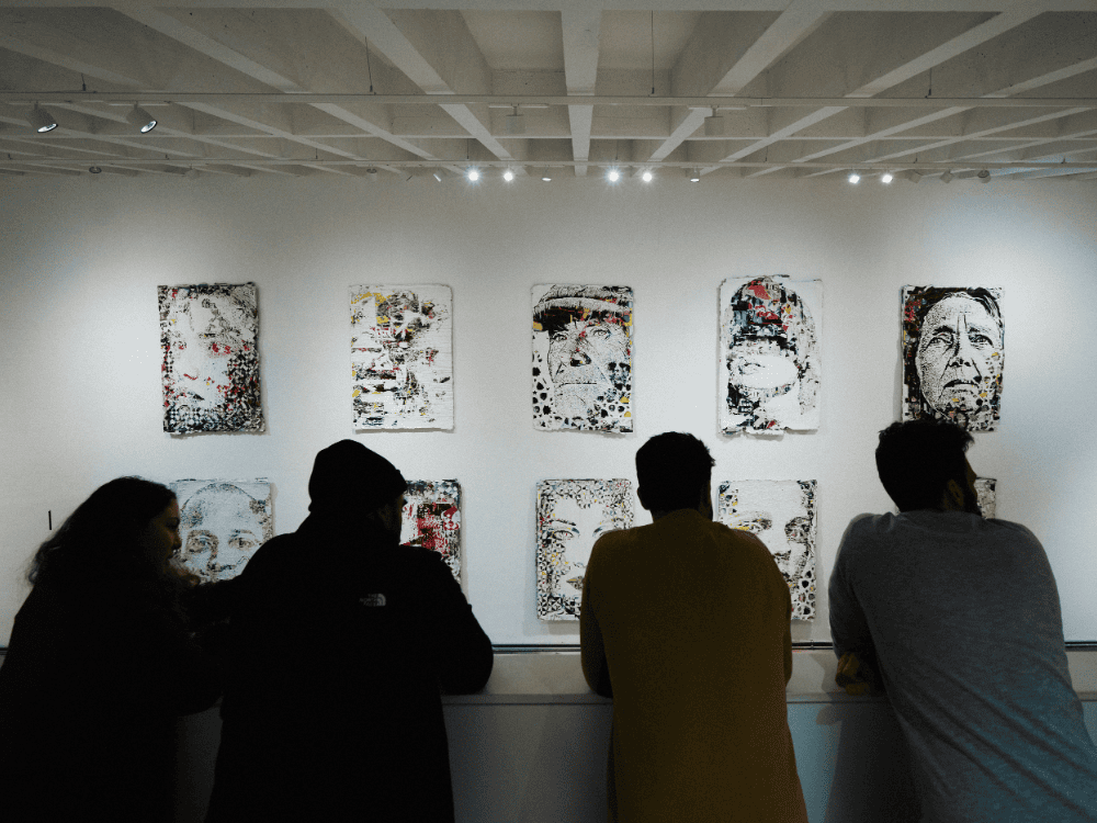 Four people stand in a gallery observing abstract portraits on a white wall. The atmosphere is contemplative, with soft lighting highlighting the artworks.