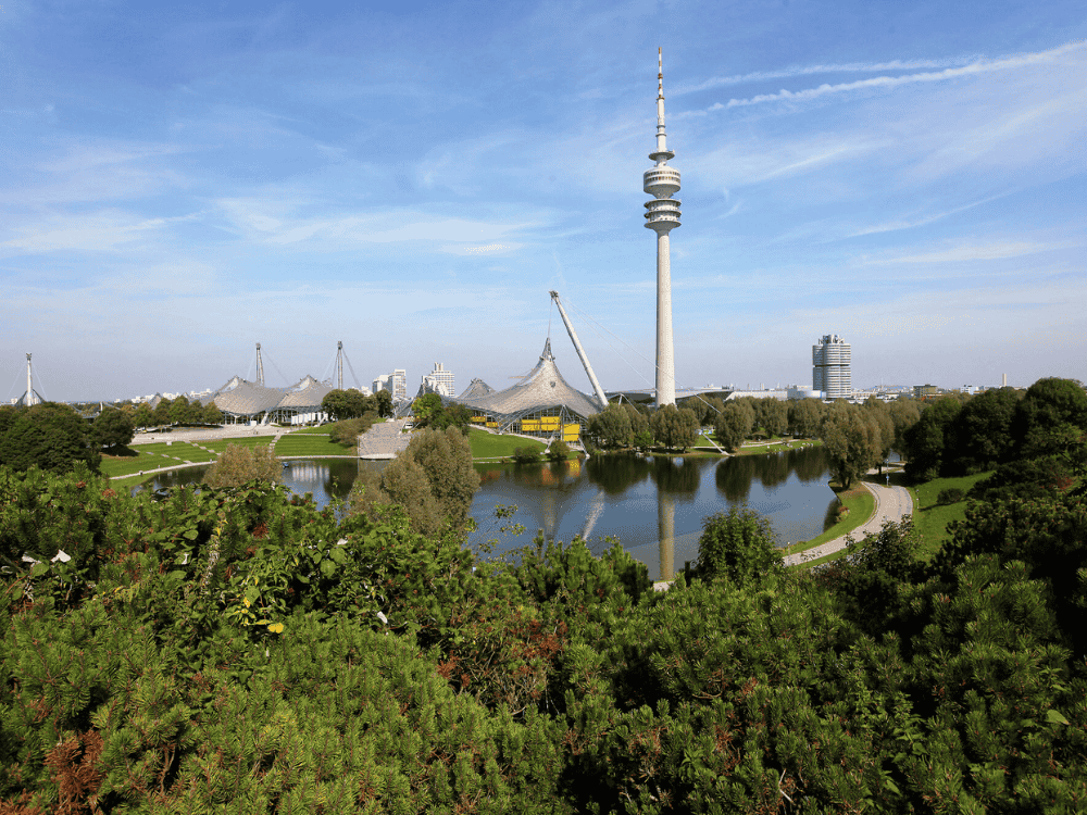 Overlooking the Olympiapark in Munich, the image shows lush greenery, a reflective lake, and the iconic Olympic Tower under a clear blue sky.