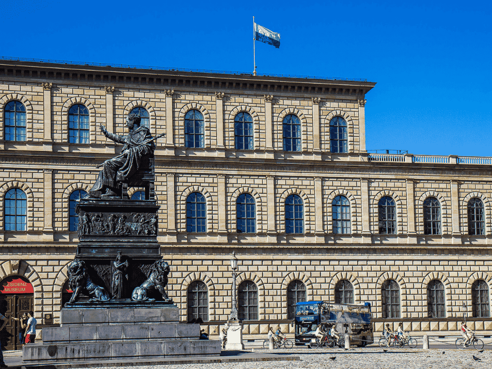 A grand statue stands before a historic building with arched windows and a flag atop. The clear sky enhances the scene's majestic feel.