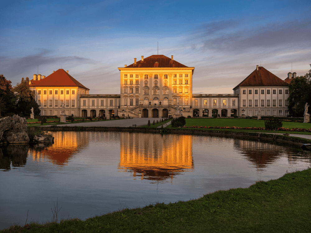 Grand palace under a vibrant sunset with a tranquil pond in the foreground reflecting the illuminated building, surrounded by lush green lawns.