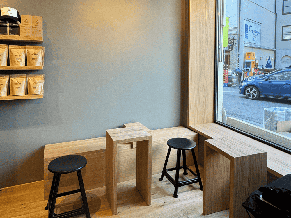 Minimalist cafe interior with wooden seating by a large window. Black stools are paired with angular wooden tables. Shelves display coffee products.