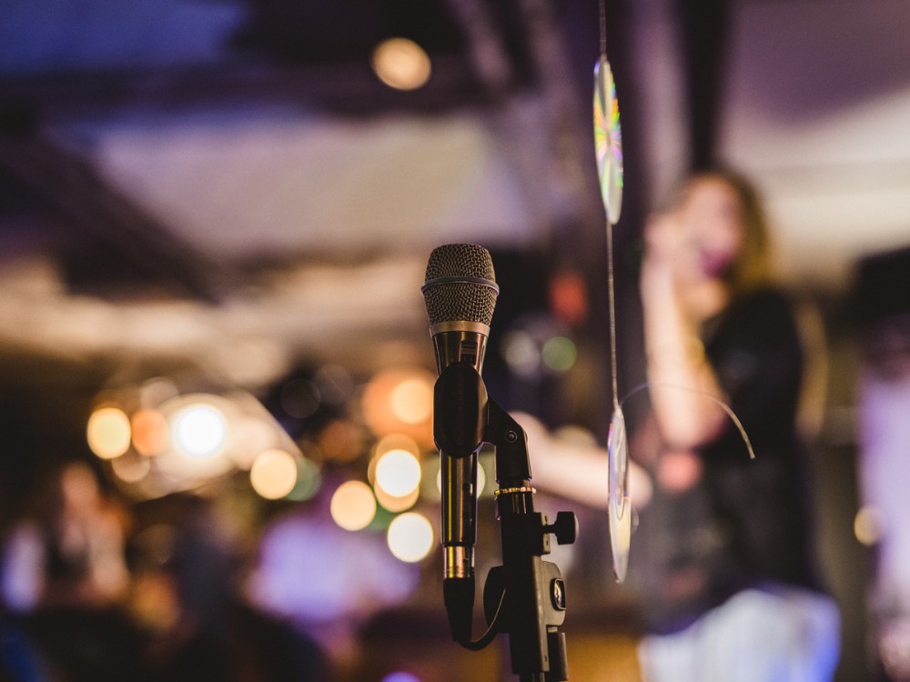 A microphone on a stand is in focus in a dimly lit venue. In the blurred background, a person appears to be singing, creating a lively atmosphere.