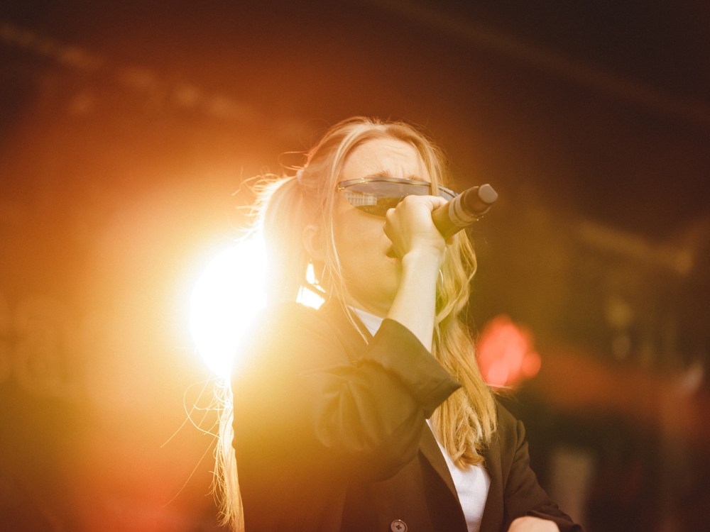 A person in a black jacket sings passionately into a microphone on stage, bathed in a warm glow from bright stage lights. The scene conveys high energy.