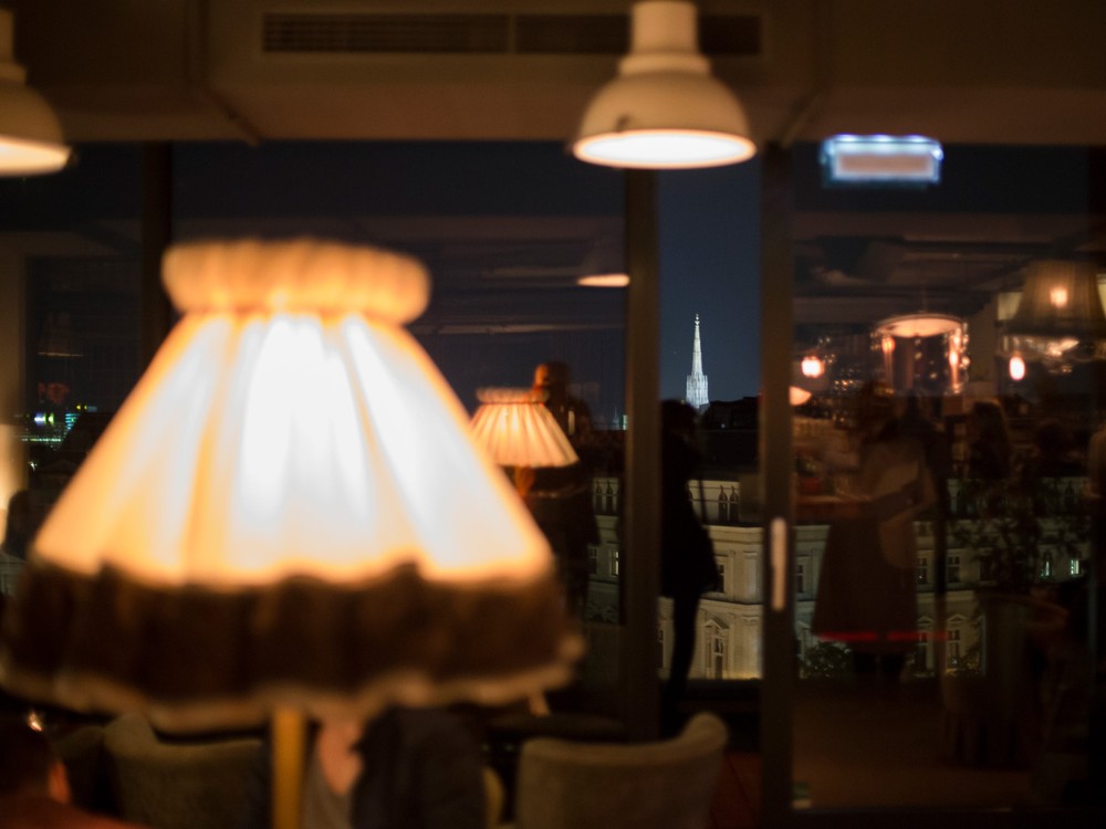 Cozy interior with a blurred lamp in the foreground, reflecting a cityscape through glass. Soft glow and night ambiance create a warm, inviting atmosphere.